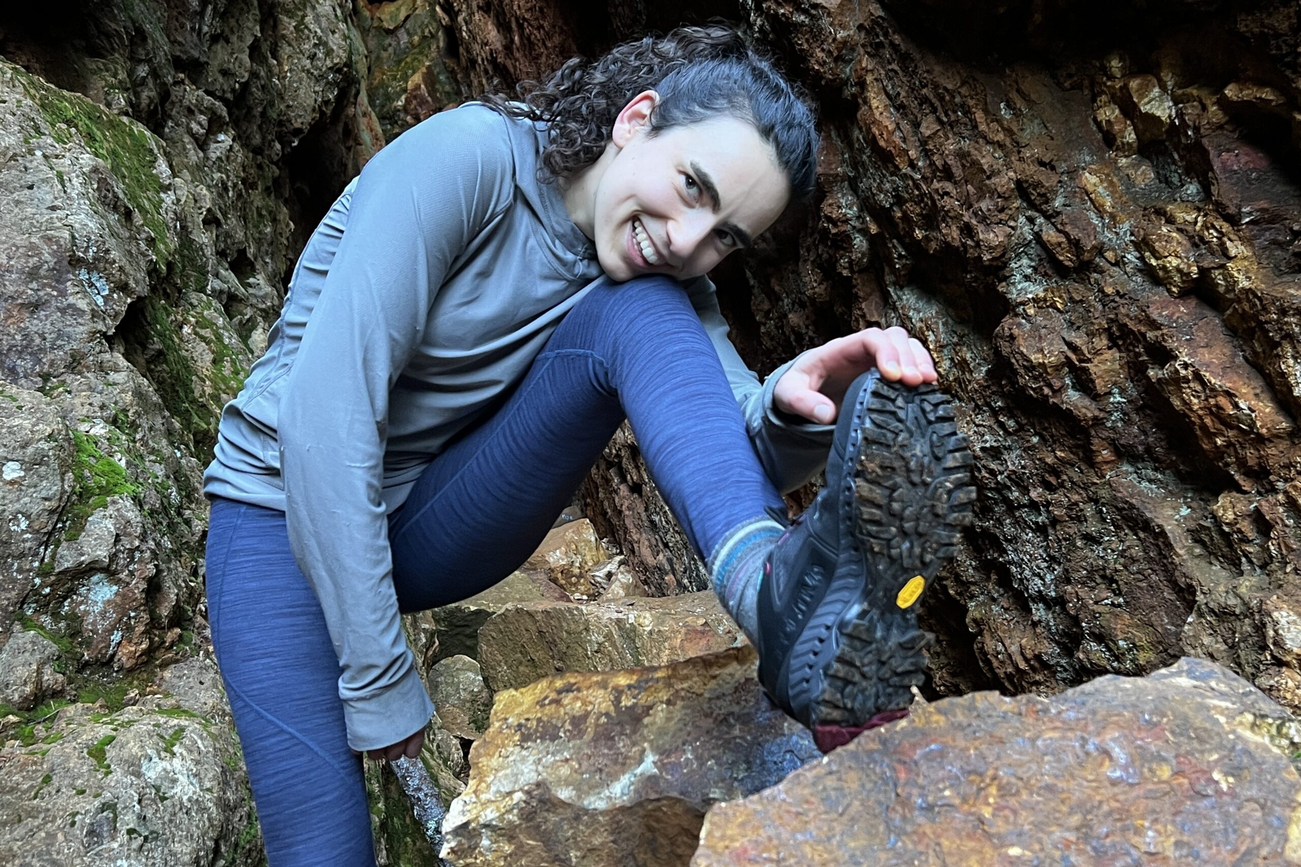 A woman pulls back a hiking shoe with her hand, as if she is stretching, to show the bottom tread.