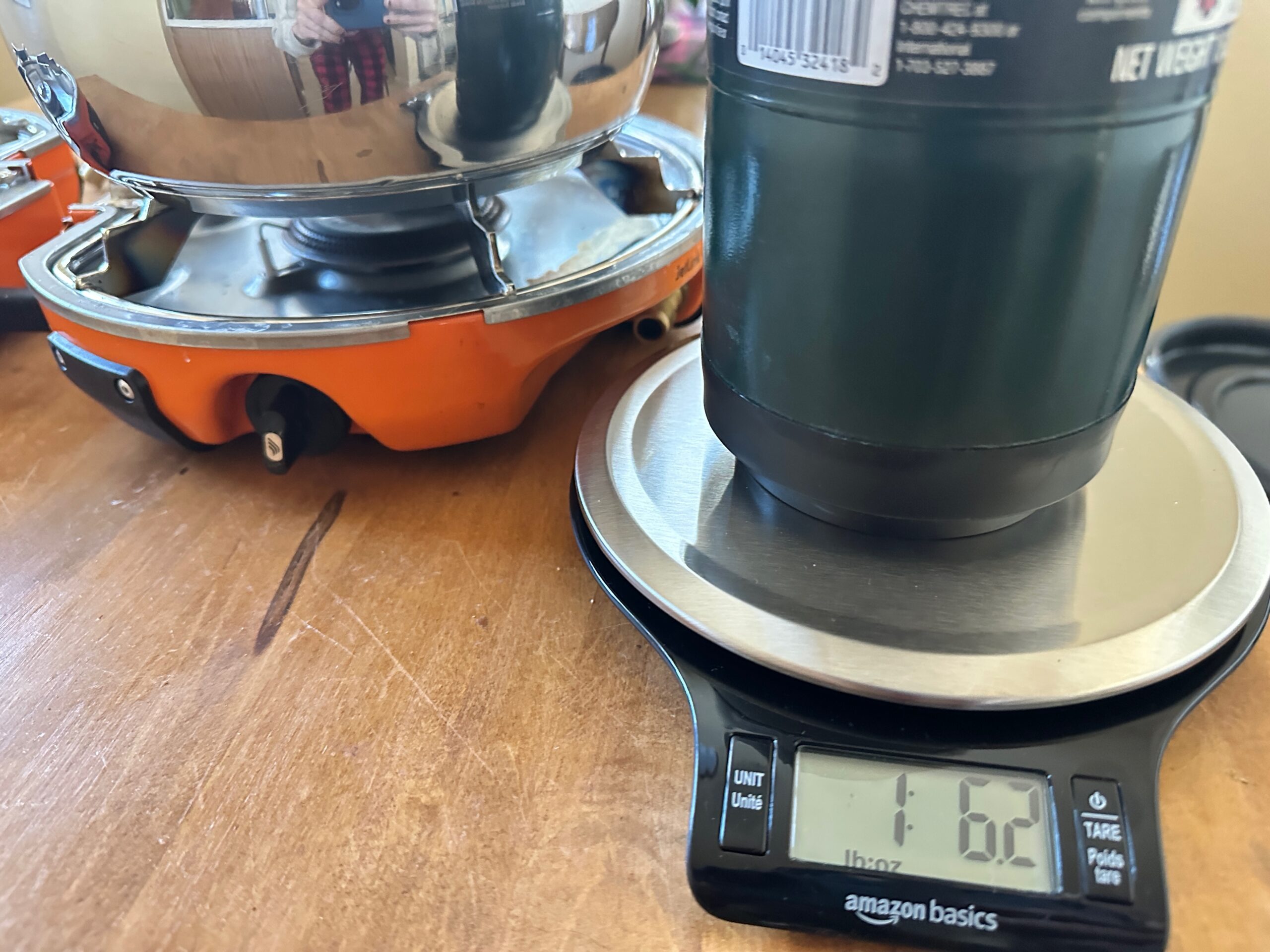 a scale with a propane canister on top with a camp stove in the background to test the amount of fuel used to boil water. 