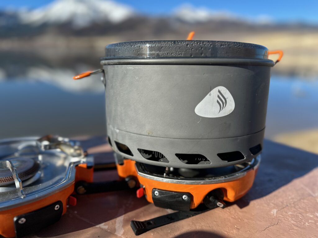 Jetboil Genesis Basecamp System at a lakeside campsite with a snowy mountain in the background