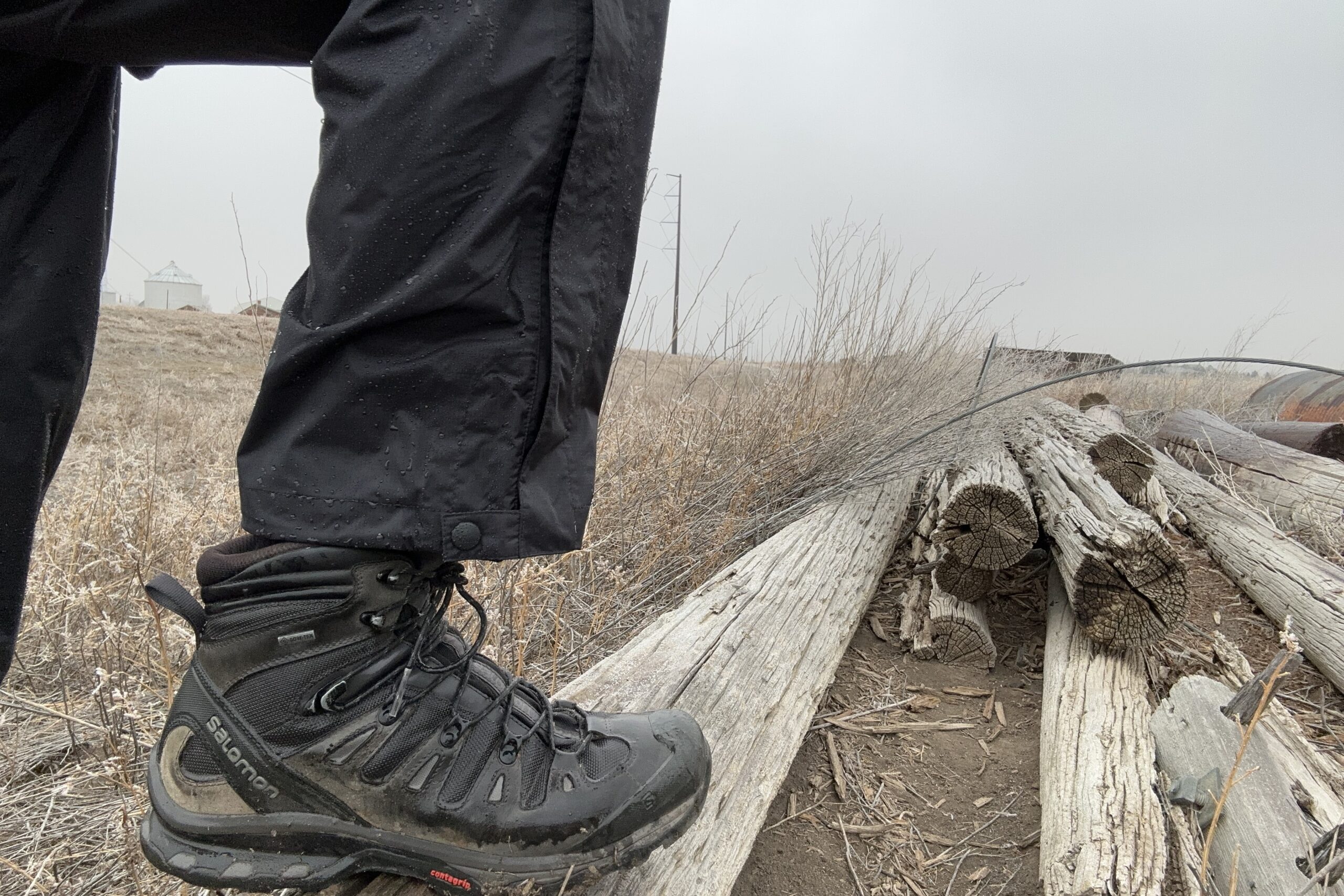 Closeup of someone wearing the black Marmot PreCip Eco rain pants, stepping up on an old fencepost on the ground.