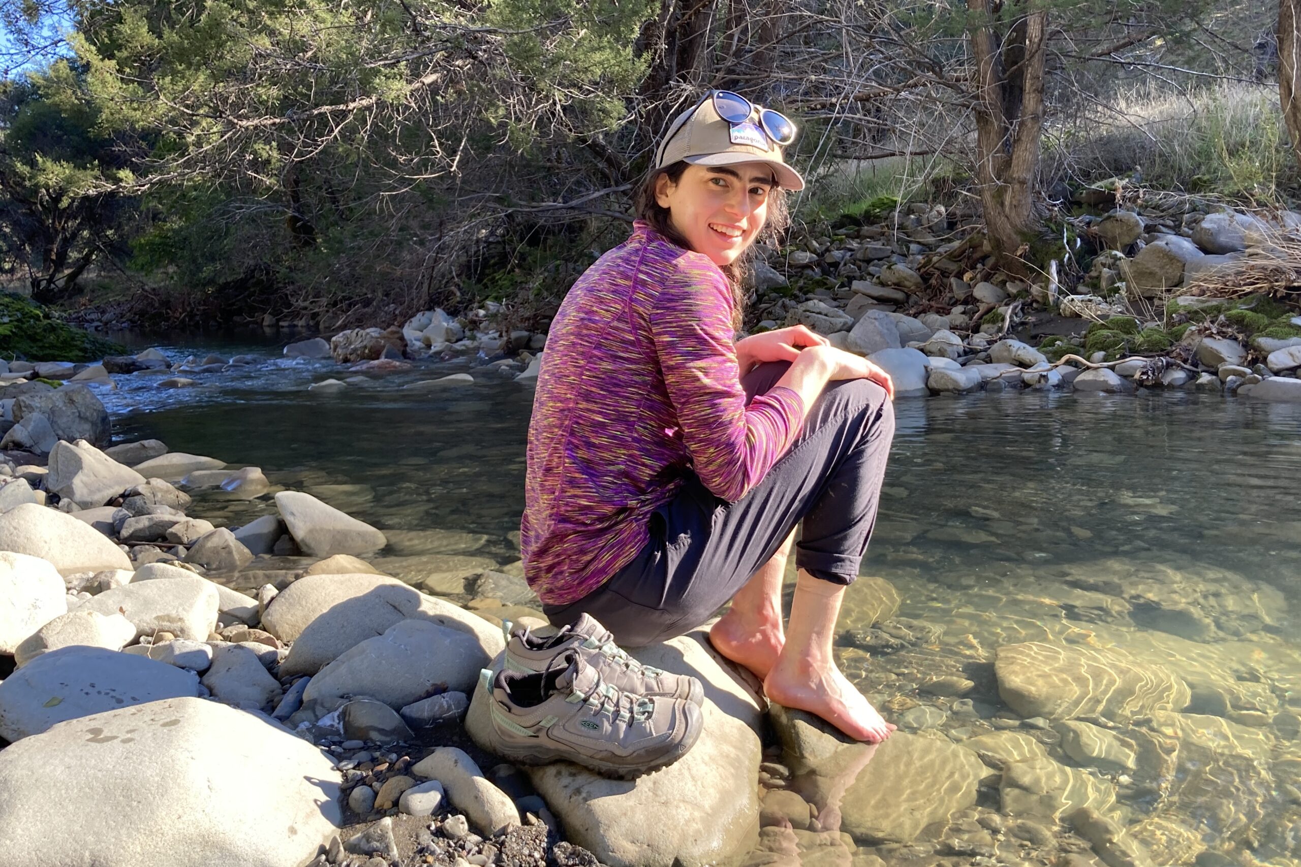 Women sitting next to a creek, soaking her feet in the water while a pair of Keen shoes sit next to her.