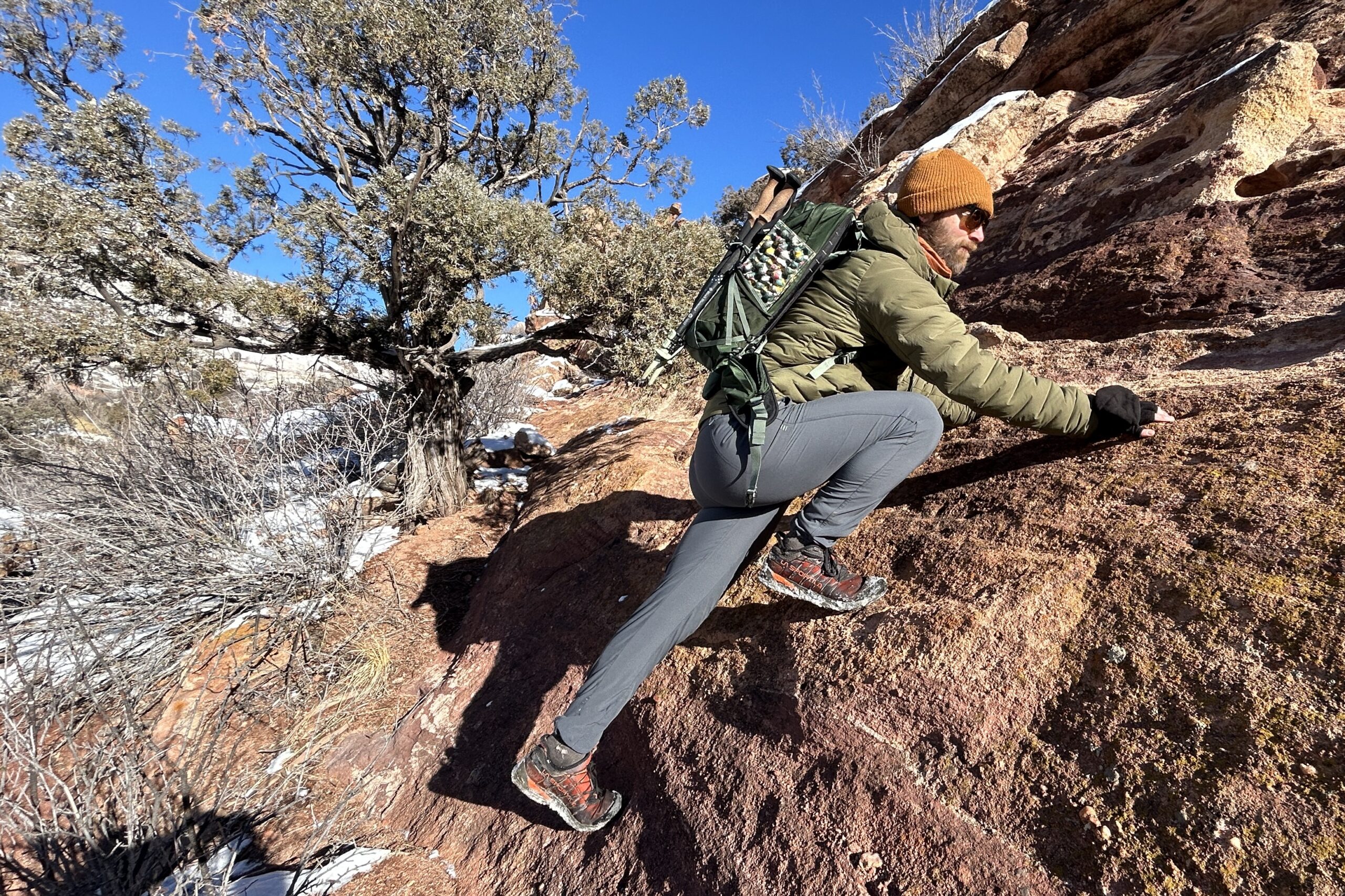 A man scrambles up a steep red rock in a snowy setting with a hat and backpack on.