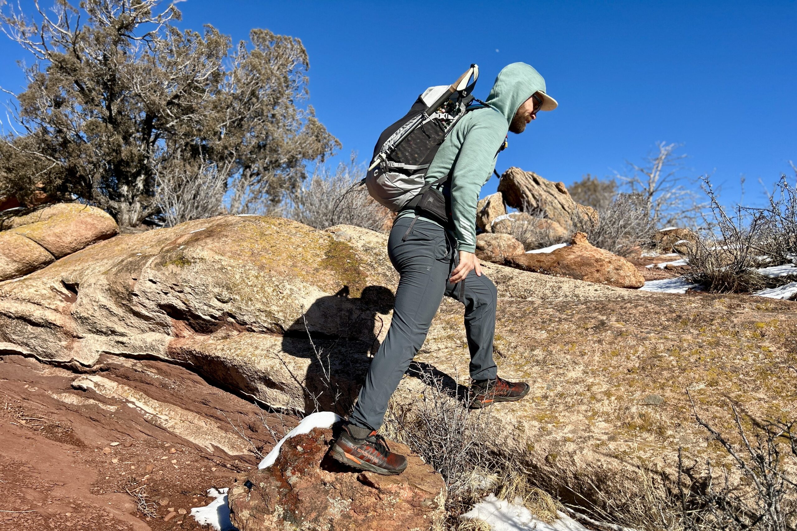 A man wearing a hoodie and a backpack hikes over sandy rocks in a desert setting.