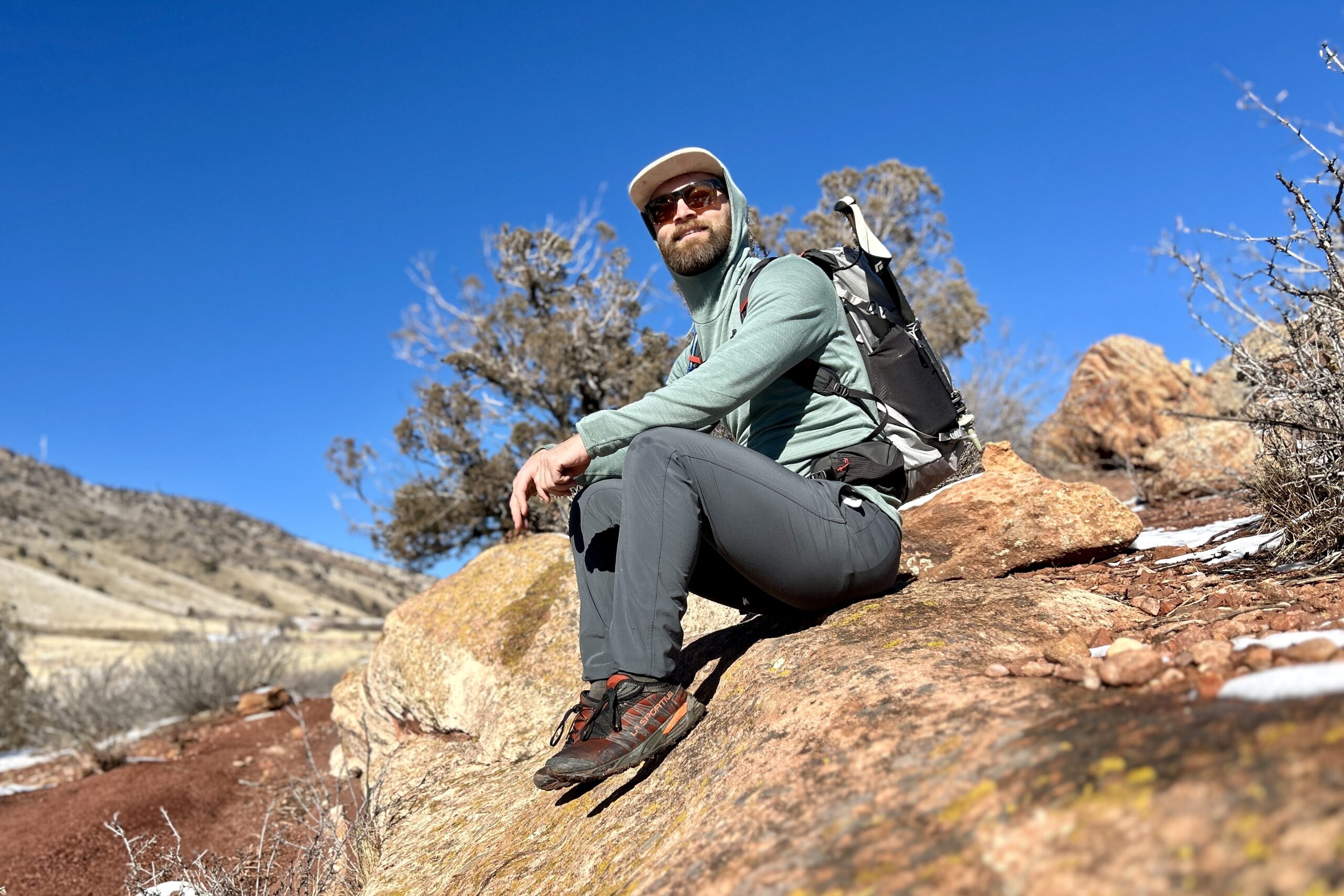 A man rests on a rock under a bluebird sky in a desert setting wearing a backpack.