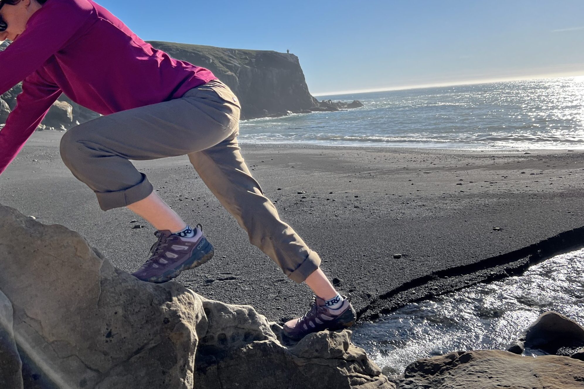 A woman is climbing a rock at the beach in a pair of Oboz hiking shoes.