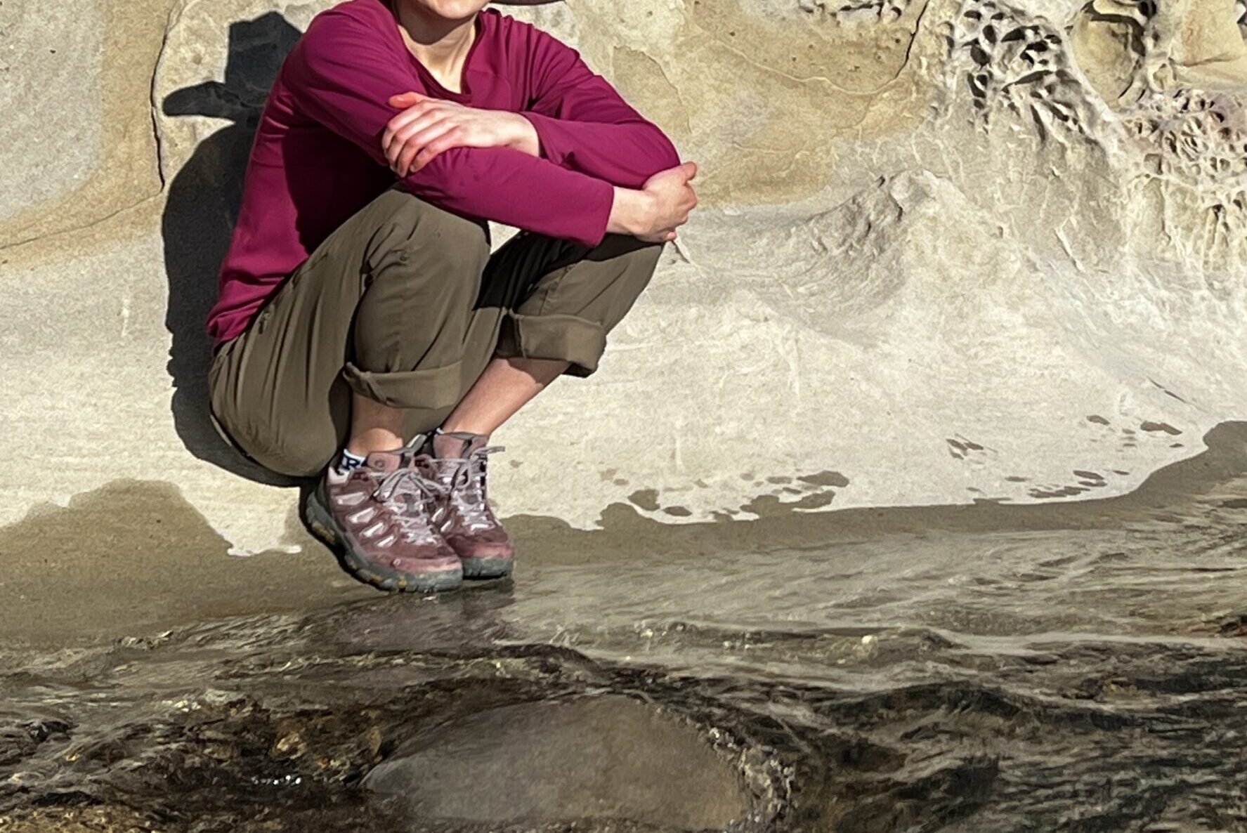 Women squatting next to a creek with a boulder in the background.