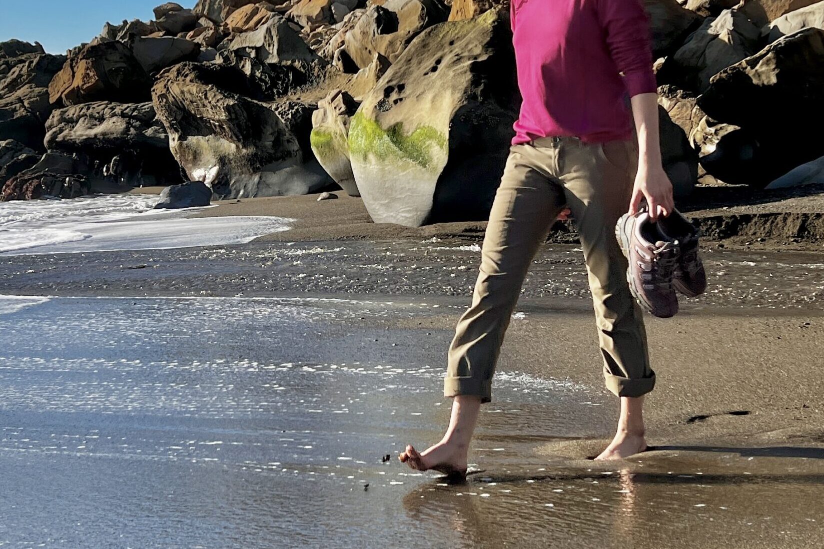 A woman carries the Oboz hiking shoes while walking barefoot on the beach.