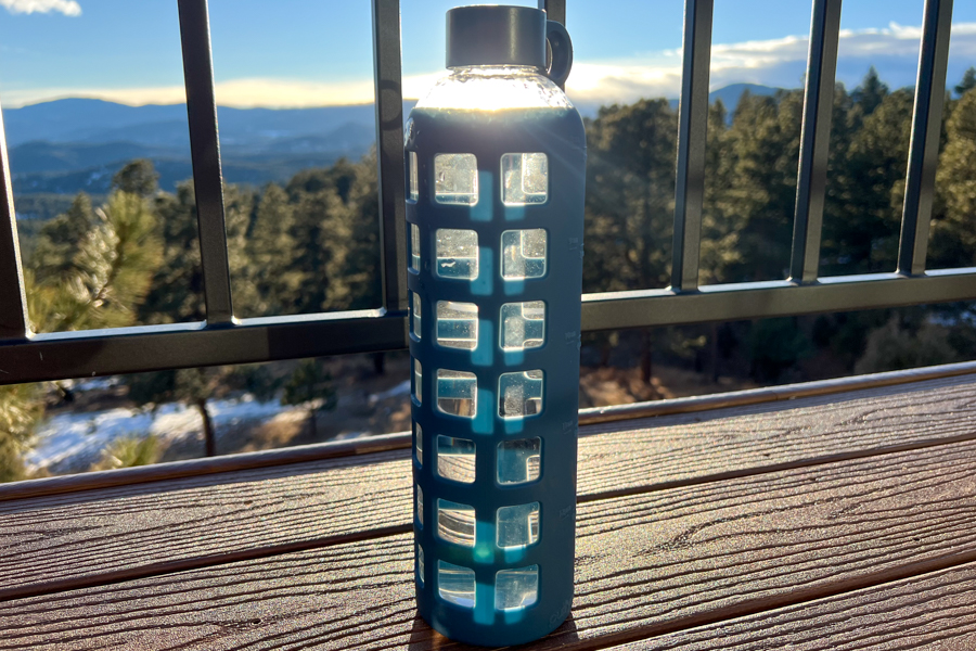 A slim glass water bottle with a geometric silicone sleeve, sitting on a wooden deck overlooking a forested view.