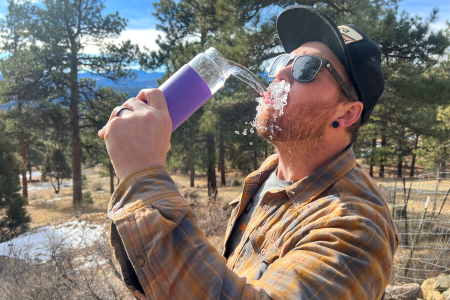 Man playfully pouring water from a purple glass water bottle into his mouth outdoors.