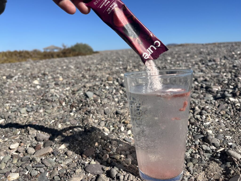 A packet of Cure electrolytes being poured into a glass on a rocky beach