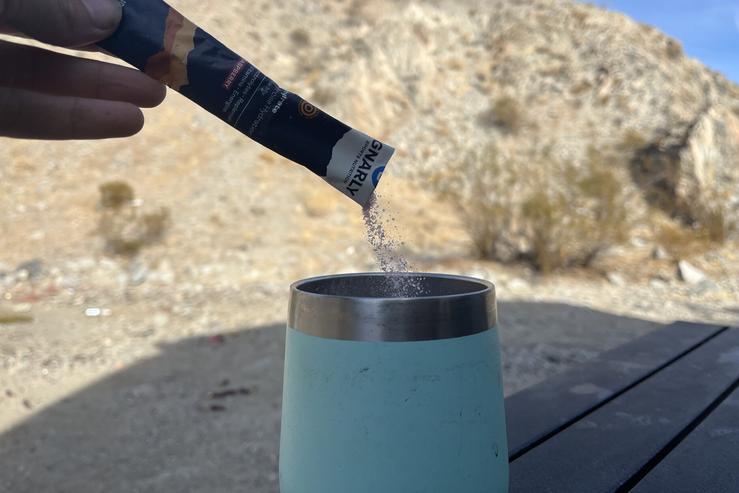 Gnarly Nutrition hydration powder being poured into a cup with mountains in background