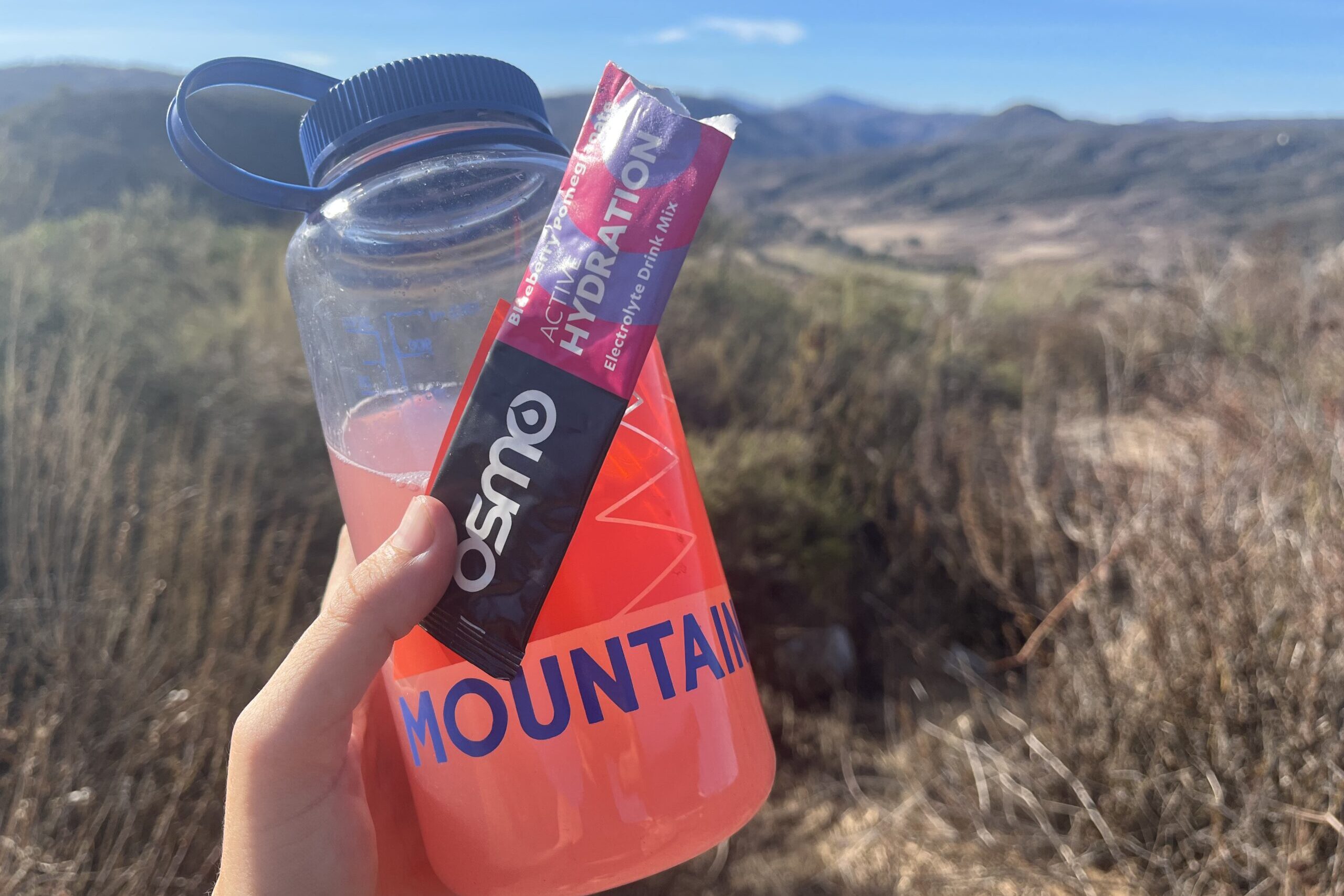 A hand holding up Osmo Active Hydration powder mixed with water in a Nalgene bottle, with mountains in background