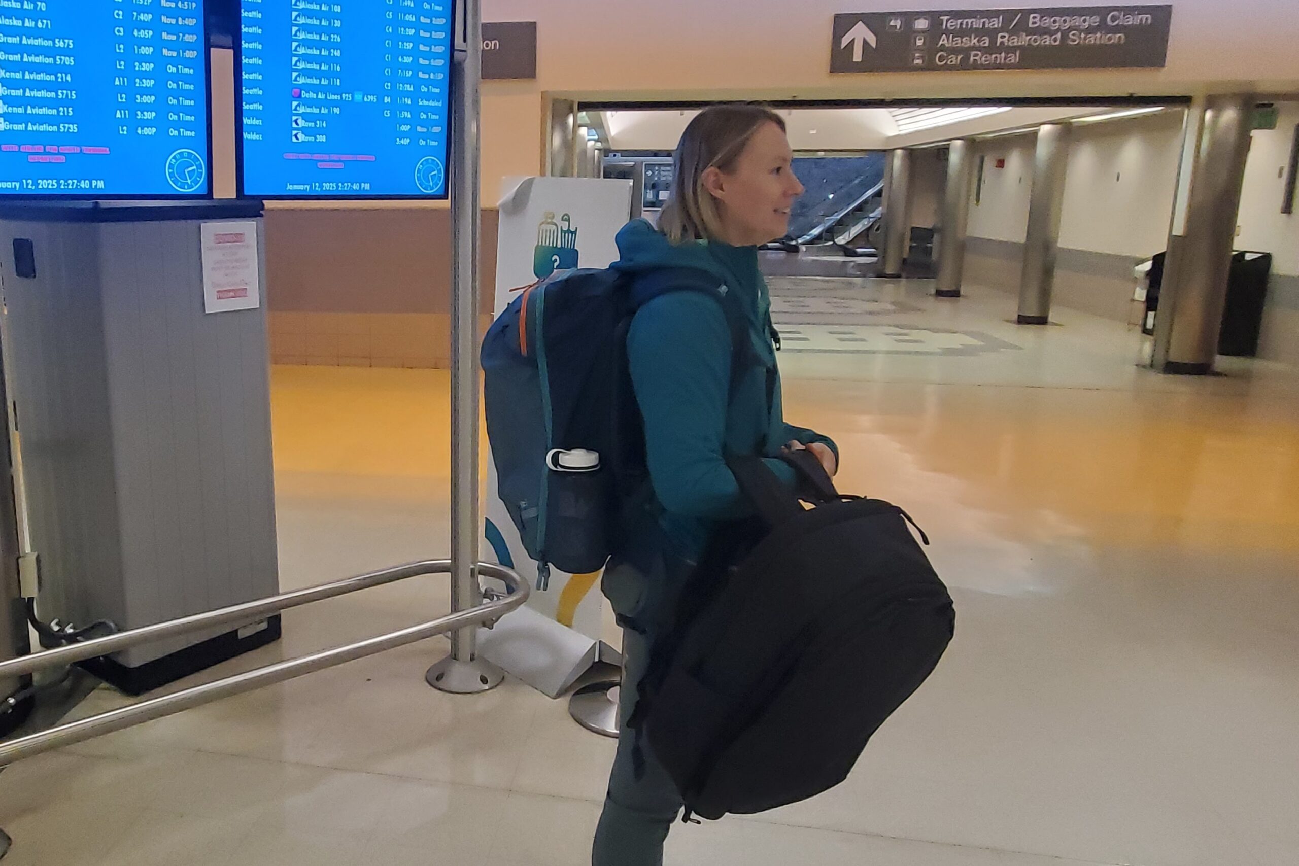 A woman stands in an airport hallway holding a black backpack.