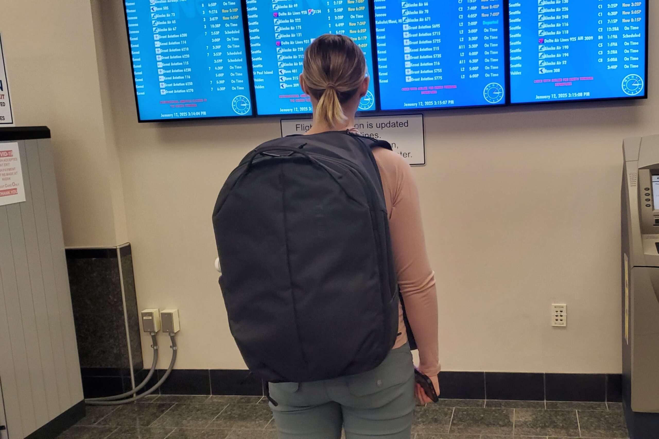 A woman stands in front of a airport monitor wearing a black backpack.