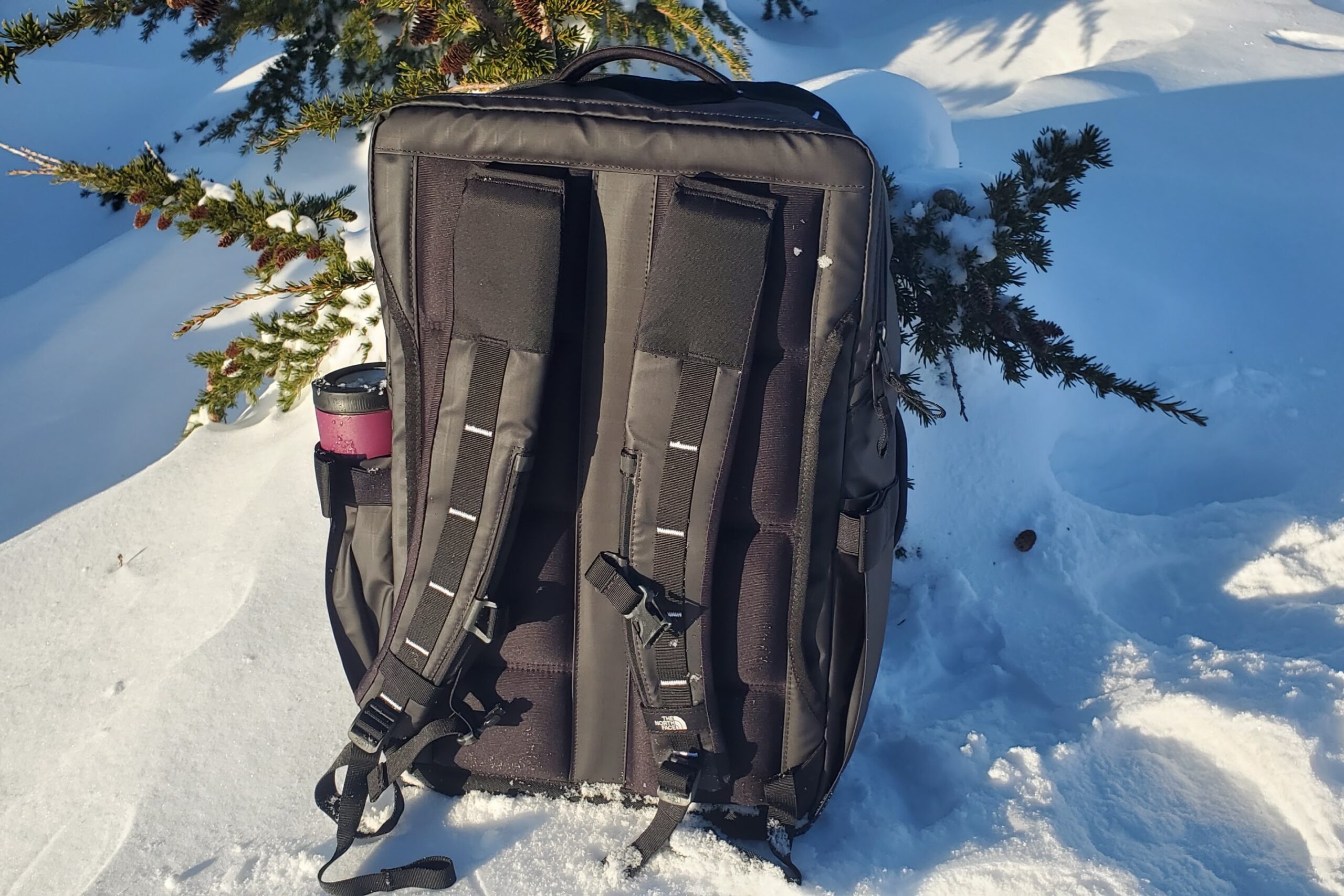 A black backpack sits in the snow in the sun.