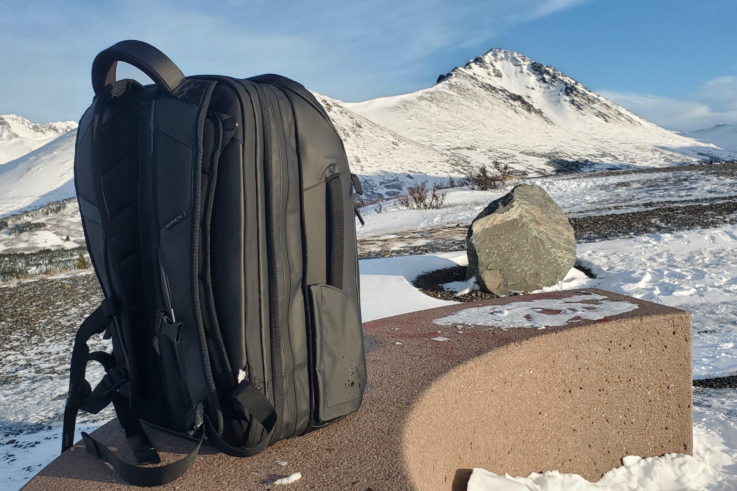 A black backpack with it's volume extended sits on a bench with a mountain view.