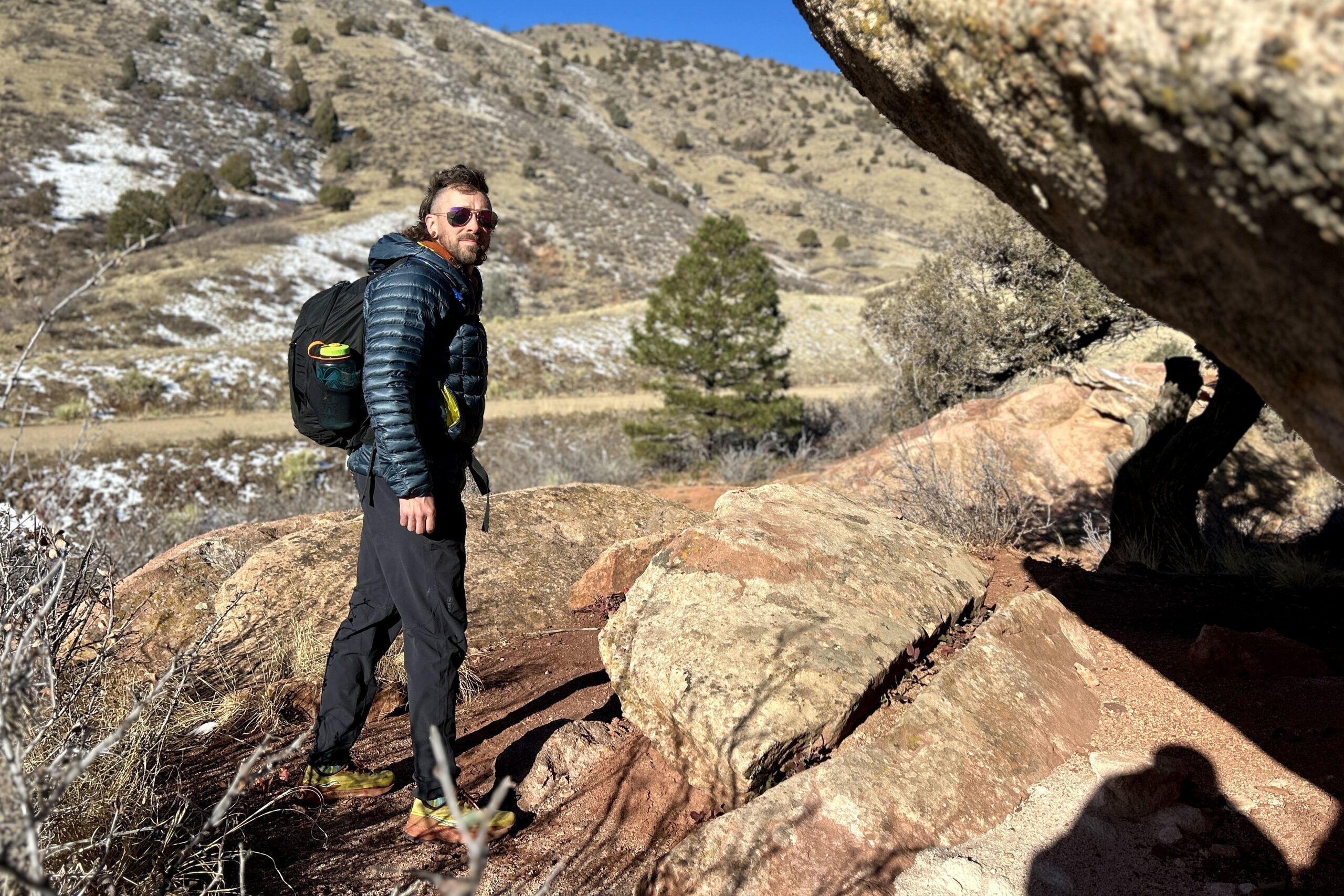 A man wearing a backpacking and a puffy jacket walks under a huge boulder.