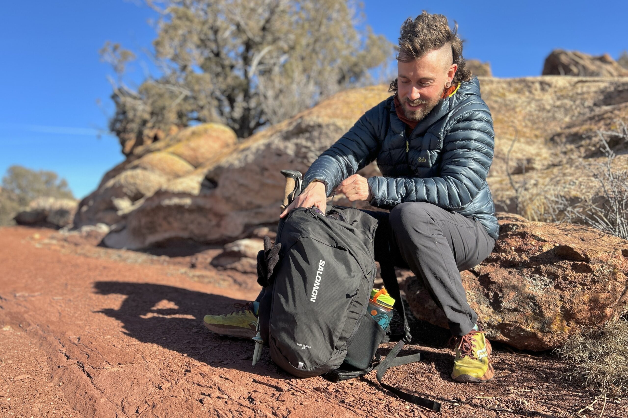 A man sits on a rock in a red desert landscape and digs through his backpack.