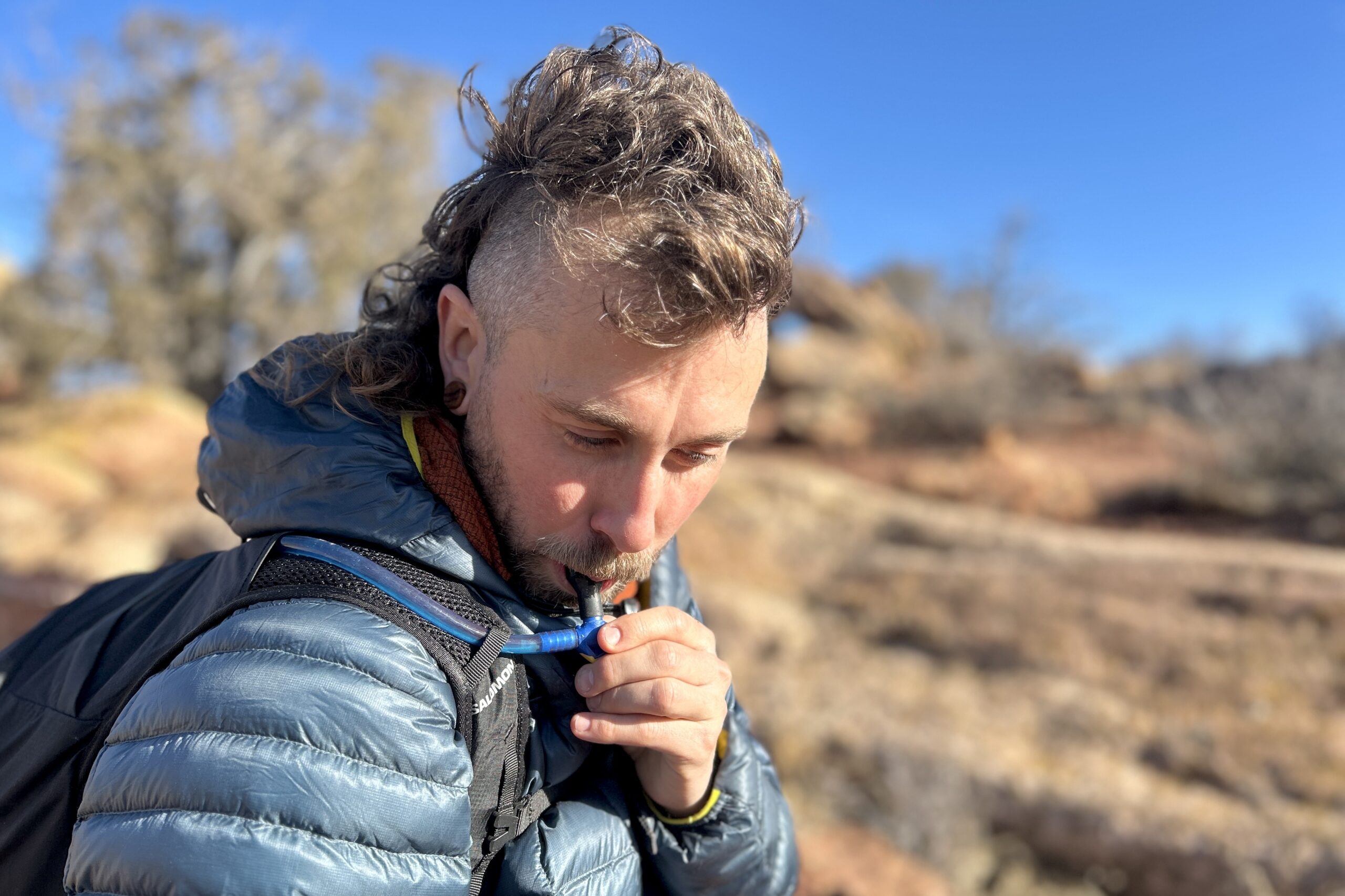 Close-up image of a man drinking water out of a hydration tube routed along his shoulder strap.