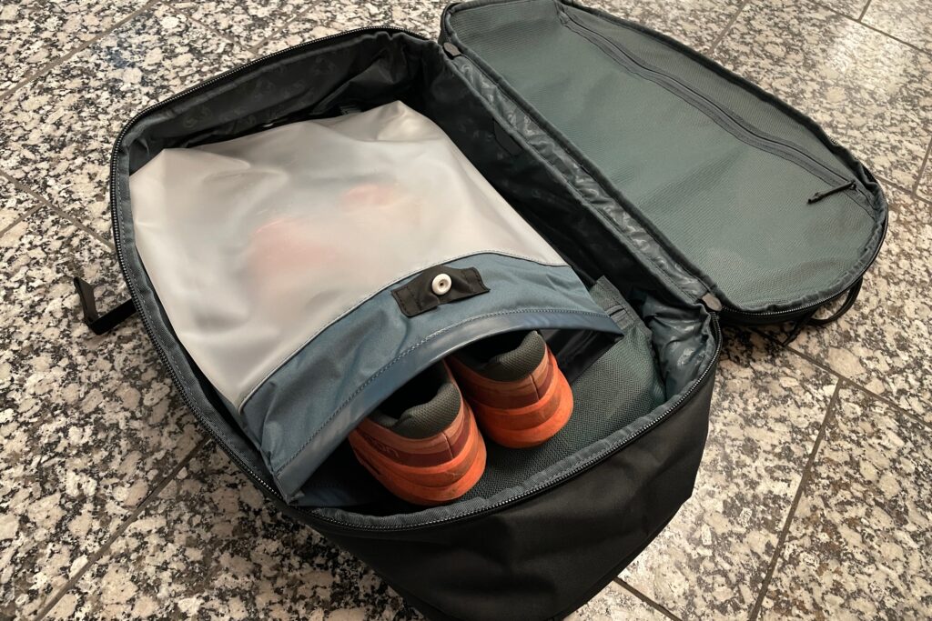 A backpack rests open on the floor with shoes in it's included dry bag.