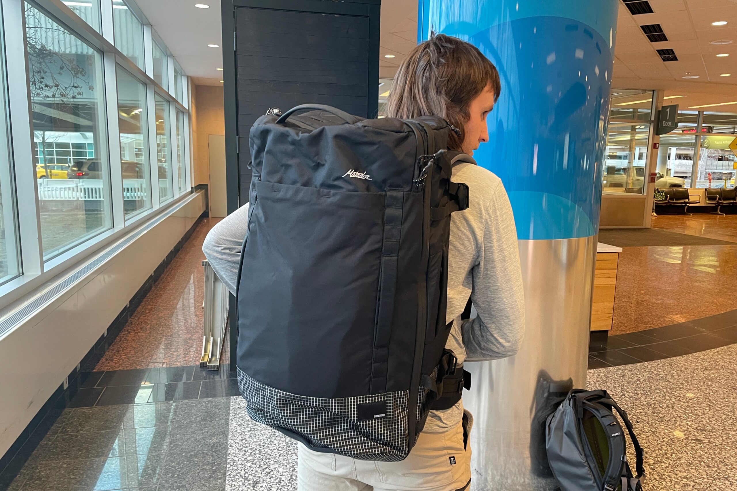 A man faces away from the camera in an airport lobby wearing a black backpack.