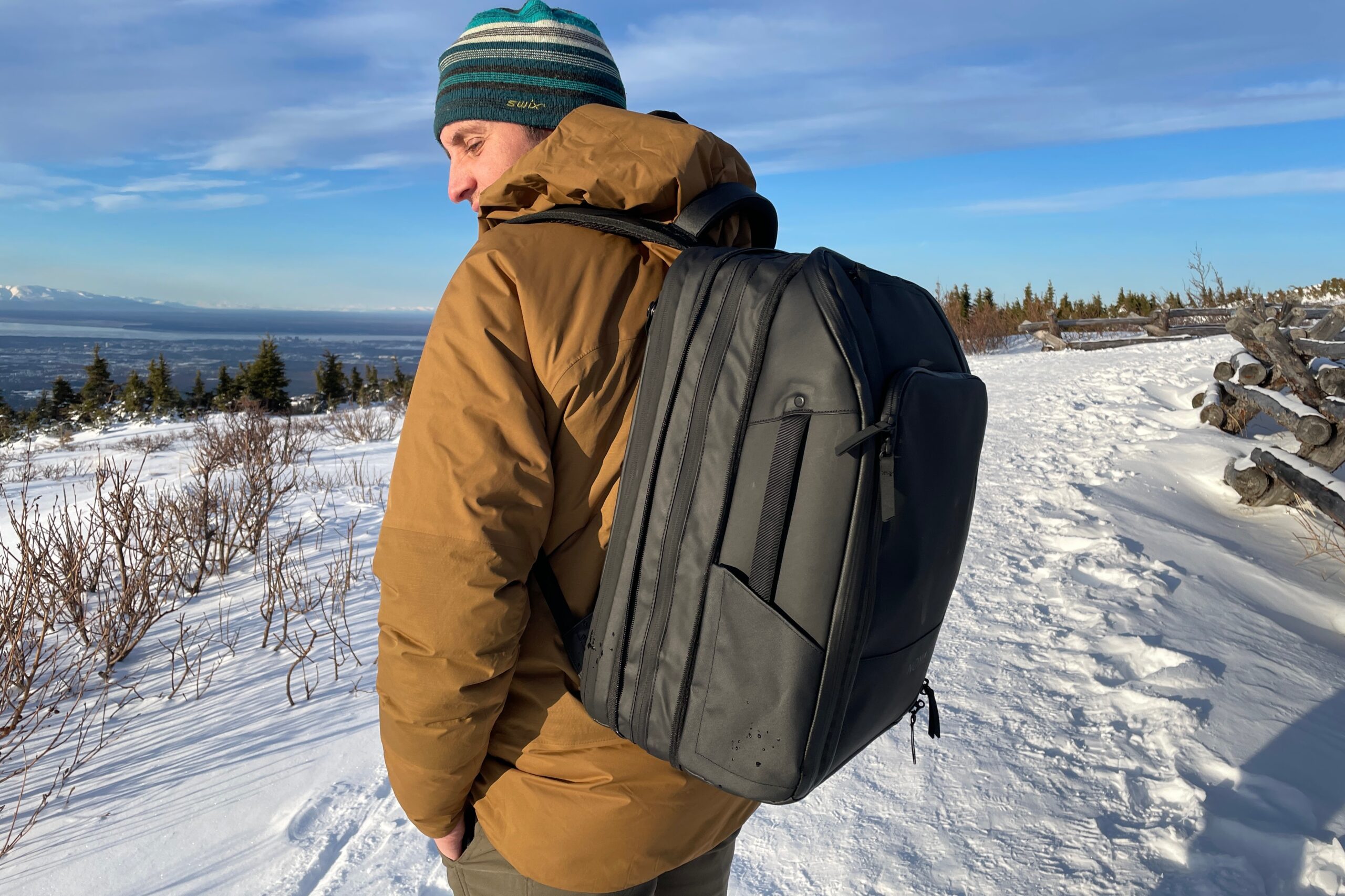 A man stands on a snowy trail wearing a black travel backpack.